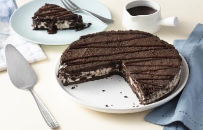 Imagem aproximada da receita de Torta de Sorvete Moça com Calda Quente, servida em um prato branco, sobre uma bancada clara decorada com um tecido azul, um pote branco com calda de chocolate e alguns utensílios