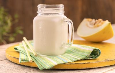 Foto em tons de amarelo da receita de smoothie de melão servida em uma jarra de vidro pequena sobre um pano listrado verde e amarelo com dois canudos ao lado. Ao fundo uma fatia grande de melão