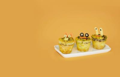 Fotografia em tons de amarelo em uma mesa e fundo amarelo escuro, um recipiente branco retangular e três mini muffins de omelete com desenhos infantis.