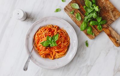 fotografia em tons de branco e vermelho de uma bancada branca vista de cima. Ao centro um prato branco com macarrão espaguete e molho de tomate, e ao lado uma tábua com folhas.