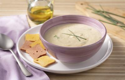 Fotografia em tons de roxo e marrom de uma bancada de madeira com um prato raso branco, sobre ele um recipiente redondo roxo com o creme de palmito. Ao lado um paninho roxo com um colher. Ao fundo uma jarra com azeite e uma tábua de madeira com cebolinha.