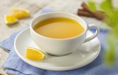 Fotografia em tons de azul, cinza e amarelo de uma bancada cinza com um paninho azul e sobre ele um prato branco redondo com uma xícara com chá de laranja. Ao fundo paus de canela e pedaços de laranja.