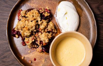 Fotografia vista de cima de um prato raso de vidro marrom, com um crumble de de frutas com creme inglês em um recipiente e chantilly de iogurte ao lado. O prato está sobre uma mesa de madeira.