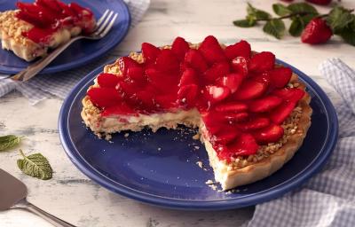 Fotografia em tons de vermelho em uma bancada de madeira de cor branca. Ao centro, um prato azul contendo a torta. Ao lado, há um pano azul quadriculado e ao fundo, um pires contendo uma fatia da torta.