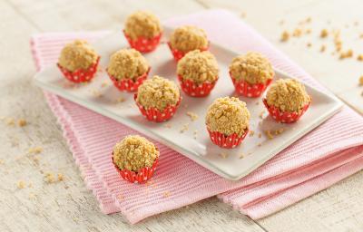 Fotografia em tons de branco e vermelho de bancada de madeira branca, pano rosa dobrado, um brigadeiro de paçoca sobre o pano,  prato retangular branco com nove brigadeiros de paçoca em forminhas vermelhas com bolinhas brancas.