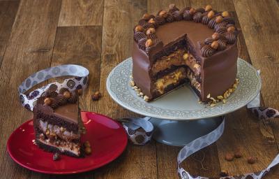 Foto da receita de bolo de chocolate com especiarias, sobre um prato cinza e sem uma fatia que está num prato vermelho do lado esquerdo. Tudo está em uma bancada de madeira decorada com uma fita de páscoa e pedaços de avelãs