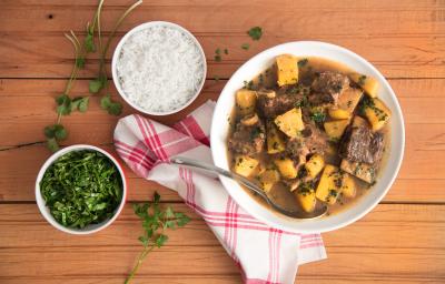 Fotografia em tons de marrom, branco, amarelo e verde de uma bancada marrom vista de cima. Contém um recipiente redondo branco com pedaços de carne e mandioca. Ao lado um pano branco e vermelho e dois potes redondos, um com arroz e o outro tempero