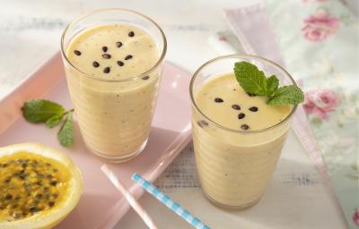 Receita de Suco Cremoso de Maracujá e Leite Moça. Observa-se dois copos com o suco decorados com hortelã e sementes de maracujá. Canudos coloridos rosa e azul e um maracujá cortado ao meio decoram a foto à esquerda.