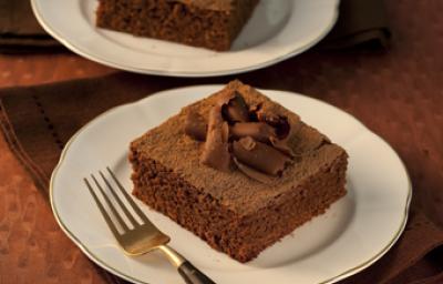 Fotografia em tons de marrom em uma mesa de madeira escura com dois pratos rasos brancos com dois pedaços do bolo de chocolate decorado com raspas de chocolate.