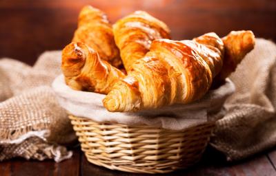 Fotografia de cestinhas de palha com um pano de tom bege. Dentro da cestinha no centro da foto, tem vários pães tipo croissant.