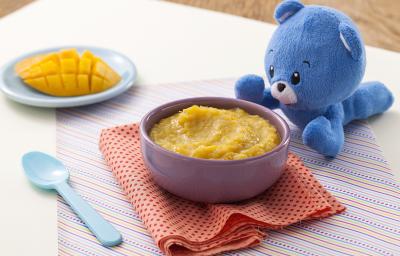 Fotografia em tons de amarelo e azul de uma bancada de madeira com um paninho listrado, um potinho roxo com o creminho de manga, ao lado uma colher azul e um ursinho de pelúcia azul. Ao fundo, um pratinho azul com um pedaço de manga cortada.