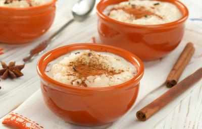 Fotografia em tons claros de três recipientes fundos e pequenos marrons sobre uma mesa clara com alguns paus de canela e cravos da índia. Dentro dos recipientes têm arroz doce com canela. Fotografia em tons claros de três recipientes fundos e pequenos marrons sobre uma mesa clara com alguns paus de canela e cravos da índia. Dentro dos recipientes têm arroz doce com canela.