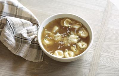 Fotografia de um recipiente branco fundo com uma sopa de capeletti, sobre uma mesa de madeira. Ao lado, um pano branco com linhas na cor cinza e preto.