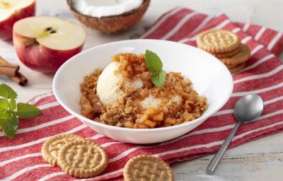 Fotografia em tons de branco e vermelho de uma bancada branca com um paninho listrado branco e vermelho, sobre ele um recipiente redondo branco com o sorvete. Ao lado uma colher e biscoitos tostines. Ao fundo uma maçã e um coco.