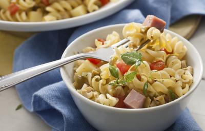 Fotografia em tons de azul em uma bancada de madeira com um pano azul ao fundo, um prato redondo fundo com a salada de macarrão tipo parafuso, com bacon e tomate.