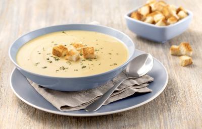 Foto da receita de Sopa de Mandioquinha com Baunilha em uma tigela azul clara, em cima de um prato azul claro e um guardanapo cinza, sobre uma tábua de madeira clara e com croutons em um pequeno pote de decoração.