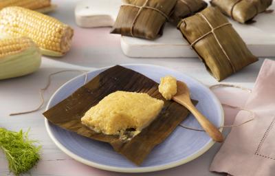 Foto da receita de Pamonha de Leite Moça, Milho Verde e Erva Doce. Observa-se a pamonha aberta em um prato de sobremesa de cerâmica com uma colherinha ao lado direito. Atrás, tem milhos na espiga e pamonhas fechadas e embrulhadas para decorar.