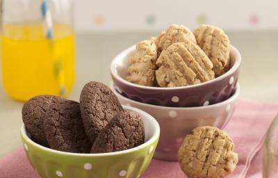 Fotografia em tons de rosa em uma bancada de madeira clara com potinhos verde, branco e marrom com bolinhas brancas em cima de um pano rosa e biscoitinhos de chocolate e aveia dentro dos potinhos.