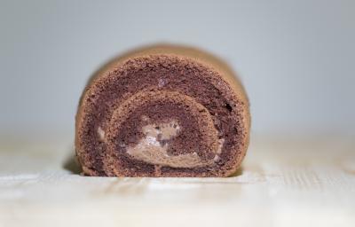 Fotografia de um bolo de rolo de chocolate, em tons de marrom, ao centro de uma mesa de madeira clara.