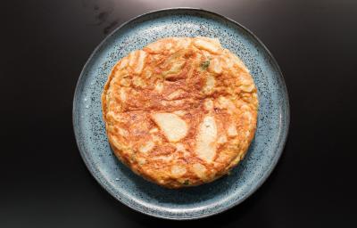 Fotografia em tons de preto com um prato azul ao centro. Dentro do prato existe uma receita de Tortilha Espanhola, uma receita feita com ovos e batatas cortadas de forma fina.