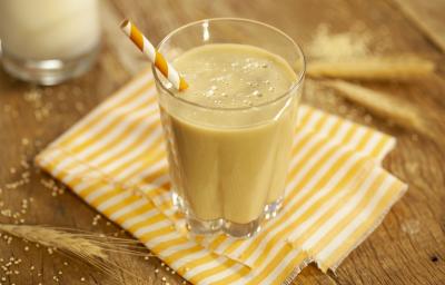 Fotografia em tons de marrom e amarelo de uma bancada de madeira com paninho listrado amarelo e branco, sobre ele um copo de vidro com a vitamina deliciosa e um canudo listrado amarelo e branco. Ao fundo ramos de trigo.