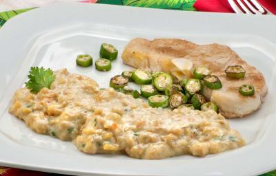 Fotografia em tons de verde em uma mesa com uma toalha verde florida, um prato branco grande com o peixe servido com quiabo e com purê de banana da terra.