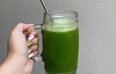 Fotografia de uma mão feminina segurando uma caneca de vidro grande com suco detox, de cor verde, com canudo de alumínio.