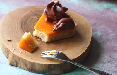 Fotografia de uma fatia de torta com damasco e mousse de chocolate por cima decorado por um bico de confeitar sobre uma tábua redonda de madeira e um garfo apoiado sobre. Ao fundo, a torta inteira sobre outra tábua de madeira redonda.