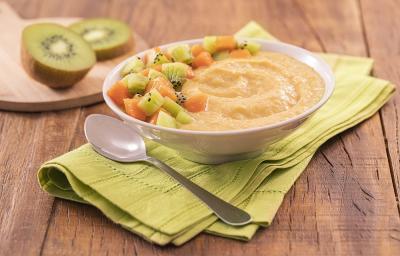 Foto em tons de verde da receita de creminho de laranja servida em um bowl de porcelana branco com uma colher prateada ao lado, sobre um pano verde claro. Ao fundo, há uma tábua de madeira com kiwis cortados em cima