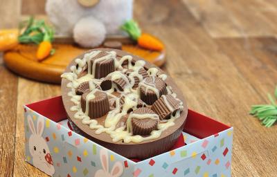 Fotografia em tons de laranja e marrom em uma bancada de madeira e uma caixa ao centro. Dentro da caixa existe um ovo de chocolate recheado com bombons e decorado com calda de chocolate branco. Ao fundo um coelho de pelúcia com cenouras.