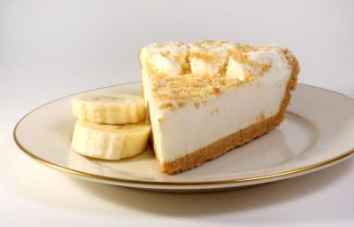 Fotografia de uma fatia de torta de banana e leite MOÇA, com uma farofinha de biscoito e rodelas de banana por cima. A torta está em um prato de vidro branco com as bordas na cor dourado, com duas rodelas de banana ao lado da torta, sobre uma mesa branca.