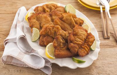 Fotografia em tons de amarelo em uma bancada de madeira clara, um pano branco com listras bege, um prato branco oval com vários bifes suínos à milanesa. Ao fundo, pratos redondos amarelos com talheres em cima deles.