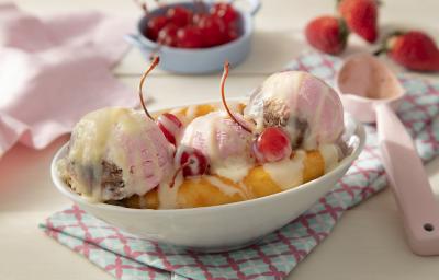 Fotografia em tons de rosa em uma bancada de madeira de cor marrom. Ao centro, uma toalha colorida e em cima, um pote contendo a banana split. Ao lado, uma colher rosa e ao fundo, um pano rosa, morangos e cerejas espalhados.