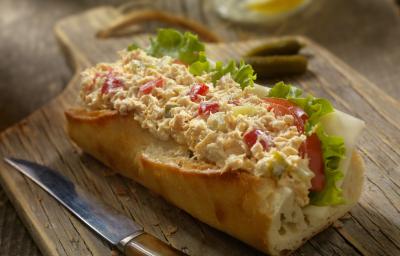 Fotografia de um sanduíche no pão francês com tomate, patê, queijo e alface, sobre uma tábua de madeira escura. Ao lado, uma faca com o cabo marrom.