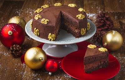 Foto da receita de Bolo Cremoso de Chocolate e Nozes, em um prato branco alto e uma fatia em um prato vermelho. Tudo está em uma bancada de madeira escura decorada com bolas de natal, pinhas e neve artificial