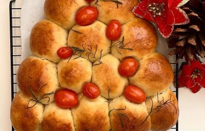 Foto da receita de Pão Caseiro Natalino. Observa-se um pão assado e dourado em formato de árvore, decorado com tomatinhos cereja