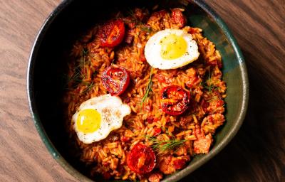 Fotografia em tons de marrom com um recipiente fundo e preto ao centro. Dentro do recipiente existe uma receita de arroz com linguiças. Em cima existe dois ovos de codorna fritos e tomates cereja tostados na frigideira.