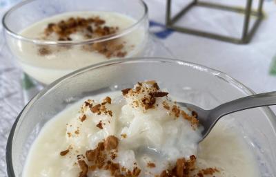 FotografiaFoto de um pote de vidro com a receita de Arroz Doce dentro, decorado com amendoins torrados. Há uma colher pegando um pouco do pote e ao fundo é possível ver a mesa decorada com uma toalha onde estão todos os itens da foto.