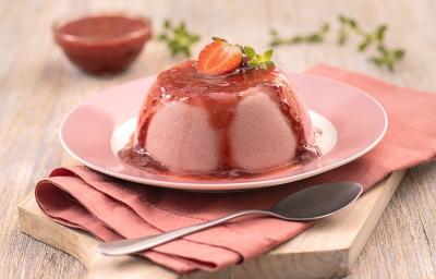 Foto em tons de vermelho da receita de flan de morango servida em um prato cor de rosa em cima de um pano vermelho sobre uma tábua de madeira retangular. Ao fundo um bowl de vidro com geleia de morango e ao lado do prato uma colher prateada.