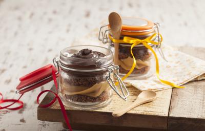 Fotografia em tons de marrom e cinza, com dois potes de vidro, um aberto e outro fechado contendo bolo fatiado, creme de chocolate e doce de leite sobre peça de madeira com colher e guardanapo ao lado, tudo sobre bancada em tons de cinza