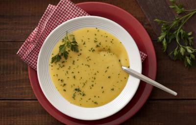 Fotografia em tons de branco, amarelo e vermelho, com prato branco ao centro contendo um creme amarelo com salsa e uma colher sobre guardanapo quadriculado em vermelho e branco e sous plat vermelho, ramo de salsa, e tudo sobre bancada de madeira rústica.