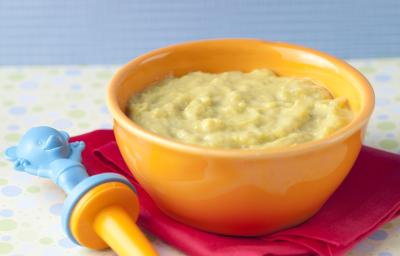 Fotografia em tons de amarelo, bordeaux e azul, com bowl amarelo contendo um creme verde claro sobre guardanapo bordeaux com uma colher infantil azul e amarela, tudo sobre toalha de bolinhas coloridas e fundo azul.