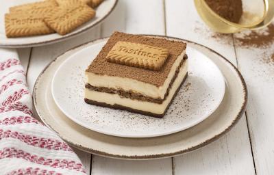 Foto da receita de Tiramisú de Tostines Especiarias. Observa-se uma fatia do Tiramisú bem alta expondo as camadas de creme e de biscoito. No topo, observa-se cacau em pó polvilhado com um Biscoito Tostines.