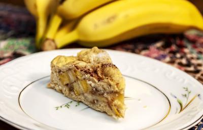 Fotografia de uma fatia de torta de banana feita com pão de forma e leite moça, que está em um prato branco com detalhes em verde e laranja. Ao fundo, um cacho de bananas.