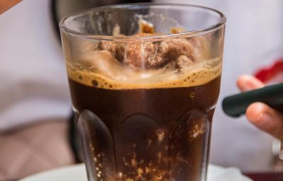 Fotografia de uma granita feita de café, leite e gelo que está em um copo de vidro, o qual está apoiado sobre um miniprato de vidro branco. Ao fundo, tem alguém segurando uma colher com a sobremesa.