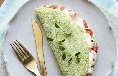 Fotografia em tons de verde em uma mesa de madeira com um pano listrado colorido em tons claro, um prato lilás redondo raso com uma tapioca verde de espinafre em cima dele.