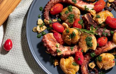 Imagem vista de cima de um prato em tom cinza com a receita de polvo, servido com batatas, tomates e algumas folhas verdes pequenas. O prato está sobre uma toalha clara e ao lado há um tábua de madeira e um tomate pequeno