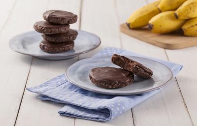 Fotografia em tons de marrom em uma bancada de madeira de cor branca. Ao centro, um pano azul claro estendido com um pires em cima contendo os cookies. Ao fundo, há mais um pires contendo cookies e uma tábua de madeira com algumas bananas.