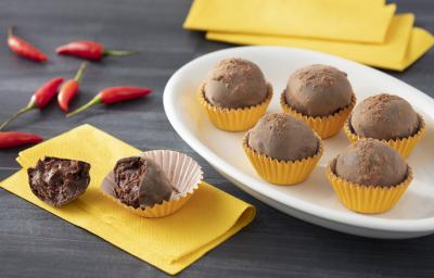 Fotografia em tons de amarelo de uma bancada cinza, sobre ela um prato oval com cinco trufas em forminhas amarelas, ao lado esquerdo um guardanapo amarelo com uma trufa cortada ao meio sobre ele. Ao fundo pimentas vermelhas.