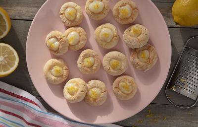 Foto da receita de biscoitos sem gluten e sem lactose com creme de limão servidos em diversas porções sobre um prato de porcelana rosa em cima de uma mesa de madeira com limões sicilianos em volta. Além disso, há um ralador e um pano